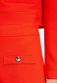 Dettaglio di una giacca rossa con un design strutturato, una texture morbida e una tasca con zip accentuata da un bottone argentato.