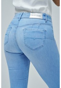 Jeans de mezclilla elástica azul claro con una cintura de media altura, que presentan bolsillos traseros con un detalle de costura curva y una etiqueta de marca en la cintura.
