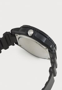 Montre noire avec un boîtier arrondi, un bracelet en caoutchouc texturé et une couronne argentée proéminente. L'arrière présente une surface métallique lisse et des vis visibles.