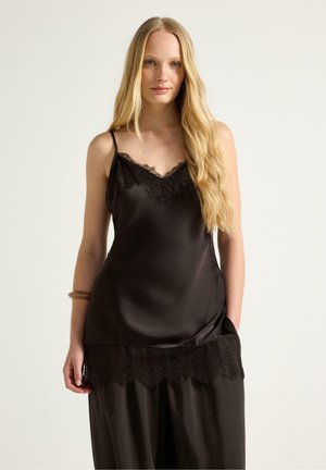 Ung kvinde med langt blondt hår iført en sort satincamisole med blonder og løse sorte bukser mod en ensfarvet baggrund.