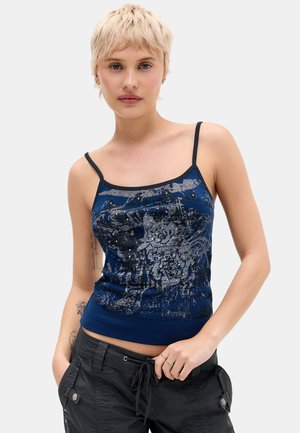 Giovane con capelli biondi corti che indossa un top blu con motivo a spalline sottili e pantaloni cargo neri, in piedi con una mano in tasca.