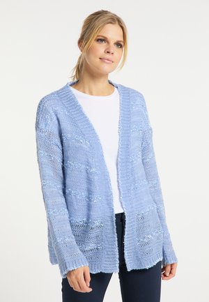 Jonge vrouw met blond haar draagt een lichtblauwe opengebreide cardigan over een wit shirt en donkere broek tegen een effen achtergrond.