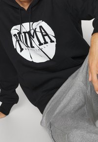 Osoba w czarnej bluzie z kapturem, z białą grafiką pękającej piłki do koszykówki i napisem "PUMA Basketball 1973", w połączeniu z szarymi spodniami.