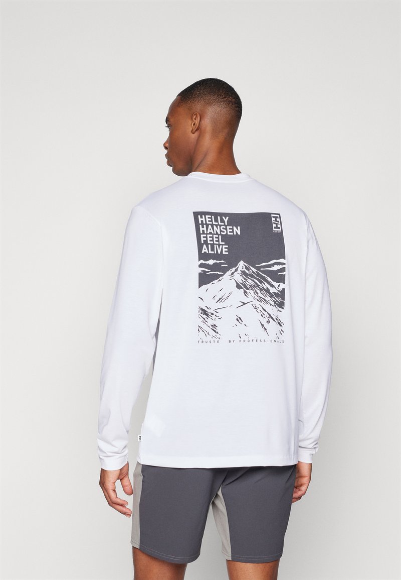 Ilga rankovė balta marškinėliai su kalno grafika pilkoje spalvoje ant nugaros, su tekstu "HELly HANSEN FEEL ALIVE." Matomos pilkos šortai.