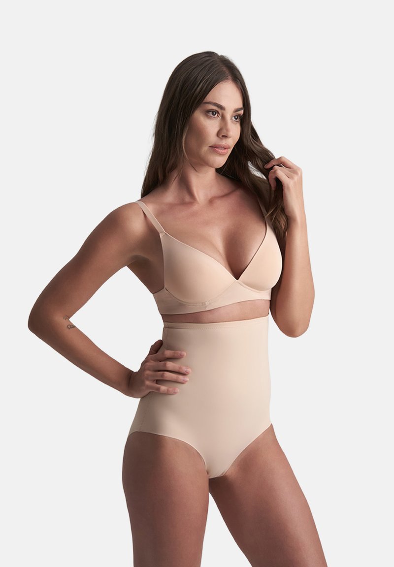 Donna con lunghi capelli castani che indossa biancheria modellante beige a vita alta e reggiseno, in posa con una mano sui fianchi e l'altra che tocca i capelli.