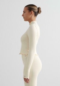 Maglia a manica lunga a coste, giallo chiaro, con collo alto e orlo a volant; abbinata a pantaloni in tinta unita a coste, con texture liscia e design aderente.