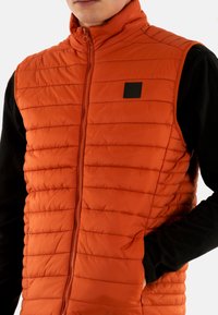 Oranje puffer vest met horizontale stiksels, hoge kraag en ritssluiting. Zwarte stoffen accenten aan de zijkant en op de borst.