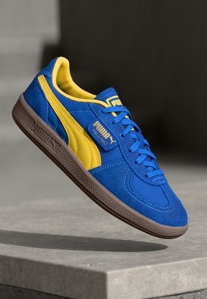 Blauwe en gele Puma Palermo sneaker met bruine zool, schuin boven een betonnen oppervlak tegen een wazige donkere achtergrond.