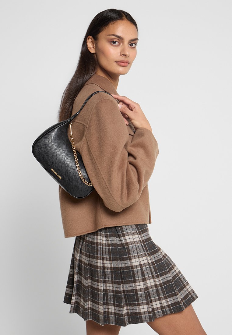 Schwarze Leder-Schultertasche mit glatter Textur, goldener Kettenriemen und geschwungenem Silhouette. Getragen mit einer braunen kurzen Jacke und einem karierten Rock.