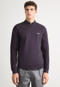 Dunkelpurpurfarbenes gestricktes Sweatshirt mit geripptem Rundhalsausschnitt, das ein kleines weißes Logo auf der Brust trägt, getragen über ein gestreiftes schwarzes Hemd.