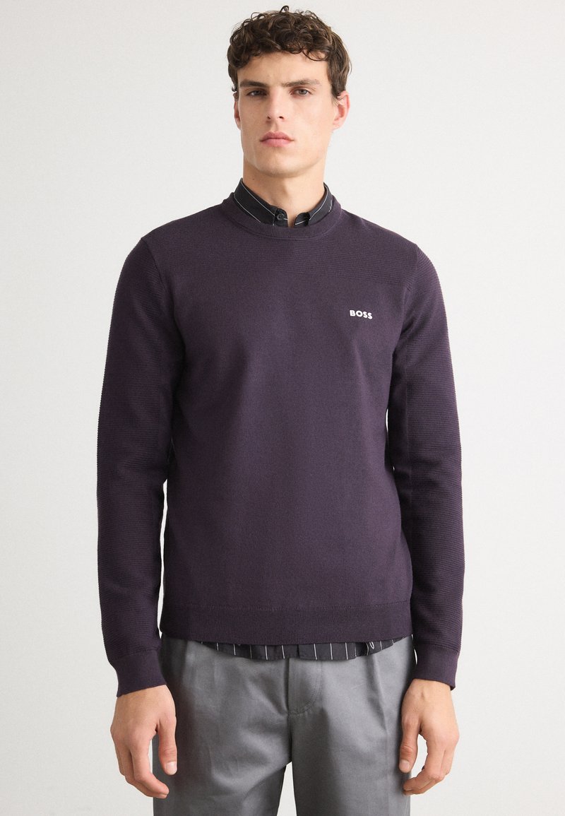 Dunkelpurpurfarbenes gestricktes Sweatshirt mit geripptem Rundhalsausschnitt, das ein kleines weißes Logo auf der Brust trägt, getragen über ein gestreiftes schwarzes Hemd.