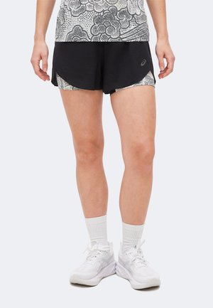 Jambes portant un short de course noir à plusieurs couches, une couche intérieure blanche à motifs, des chaussettes blanches montantes et des chaussures de sport blanches, debout sur un fond clair.