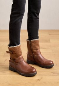 Braune Knöchelstiefel mit einer glatten Lederoberfläche und Wildlederelementen. Verfügt über ein Futter aus Kunstfell, Riemen-Details und einen Seitenreißverschluss. Robuste Sohle.