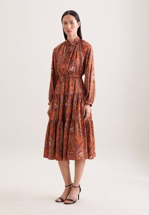 Frau trägt ein langärmeliges, knöchellanges orangefarbenes Kleid mit floralen Paisley-Muster und schwarze High-Heel-Sandalen mit Knöchelriemen.