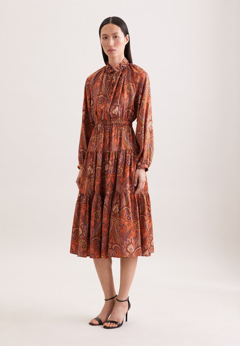 Femme portant une robe orange à manches longues et longueur cheville avec un motif floral cachemire, ainsi que des sandales à talons hauts noires avec bride à la cheville.