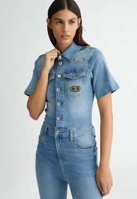 Hellblauer Jeans-Overall, kurze Ärmel, Knopfleiste vorne, Brusttasche mit Logo und figurbetonte Passform mit schmal zulaufendem Bein.
