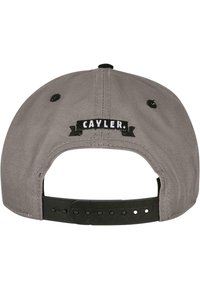 Cappellino snapback in tessuto grigio con un accento in nastro nero con il testo "CAYLER." Include una fibbia regolabile in plastica sul retro.
