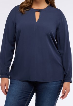 Nainen pukeutuneena tummansiniseen, pitkähihainen paitapuseroon, jossa on avainreikäkaulus, yhdistettynä tummansinisiin farkkuihin, seisoo rennosti.