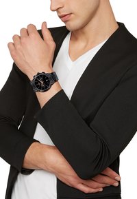 Montre chronographe noire avec un bracelet noir texturé, grand cadran noir, accents argentés et affichage de la date. Portée au poignet contre un blazer sombre.