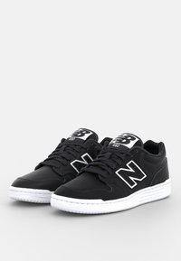 Baskets basses New Balance noires avec semelles blanches, lacets noirs et un logo blanc "N" bien visible sur les côtés et la languette.