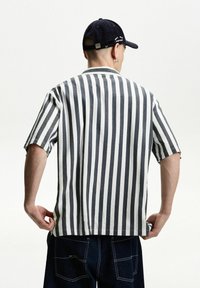 Short-sleeve shirt featuring vertical black and white stripes, relaxed fit, and a lightweight fabric; paired with dark denim pants.