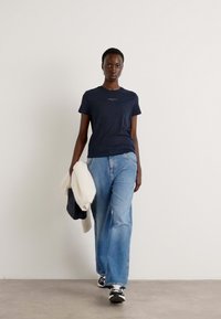 T-shirt blu navy a maniche corte con logo, abbinato a jeans a gamba larga azzurri, mentre tiene un maglione bianco e una borsa scura, indossato con sneakers nere.