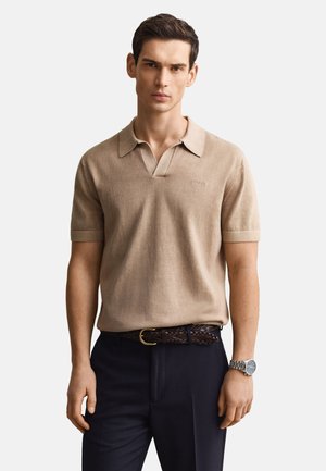 Junger Mann, der ein beige kurzärmeliges, kragenbewehrtes Strickhemd, dunkle Hosen, einen geflochtenen braunen Gürtel und eine silberne Armbanduhr trägt.