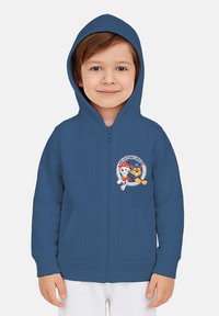 Bambino che indossa un hoodie blu con il logo del cartone "Adventure Awaits", sorridente e in piedi contro uno sfondo chiaro e semplice.