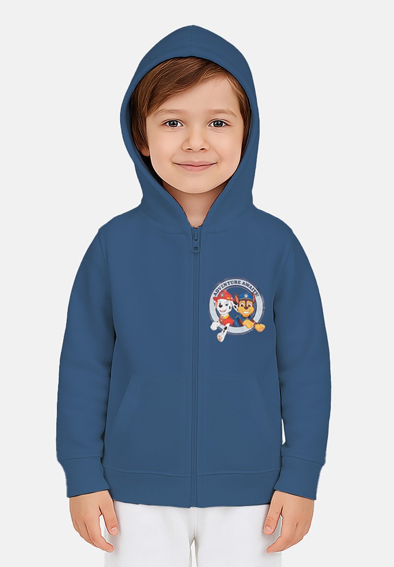 Bambino che indossa un hoodie blu con il logo del cartone "Adventure Awaits", sorridente e in piedi contro uno sfondo chiaro e semplice.