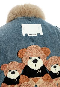 Chaqueta de mezclilla con cuello de piel sintética, que presenta tres aplicaciones de osito de peluche texturizadas vistiendo camisetas negras y una etiqueta blanca en el cuello.