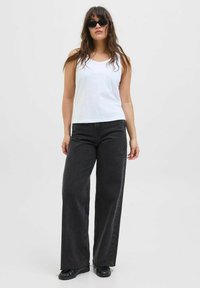 Top sin mangas blanco combinado con jeans negros de pierna ancha y cintura alta, mostrando un ajuste relajado y detalles de dobladillo deshilachado. Zapatos negros completan el look.