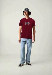 Maroon cotton t-shirt with "GAP" logo, paired with light blue jeans and black sneakers. Gray cap with text accents. Simple, casual outfit.
