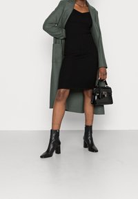 Robe noire côtelée avec une silhouette ajustée, associée à un long manteau vert, des bottines noires à talons blocs et un petit sac à main noir.