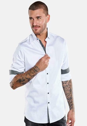Man with short hair and tattoos wears white shirt with black buttons and patterned trim, adjusting collar while looking sideways.