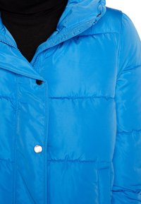 Veste rembourrée bleue avec capuche, présentant une texture lisse et brillante, des boutons pression et une poche à l'avant. Des panneaux cousus ajoutent de la dimension.