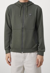 Felpa verde con zip realizzata in morbido tessuto, caratterizzata da un logo nero sul petto e due tasche laterali con zip con dettagli in nero.