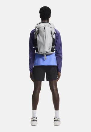 Zaino grigio con dettagli neri, spallacci regolabili e cerniera frontale. Indossato sopra una giacca blu e pantaloncini neri, visto da dietro.