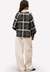 Vrouw in een zwart, grijs en beige geruite lange mouwen shirt met beige wijdvallende broek, staand en met de rug naar de camera tegen een effen achtergrond.