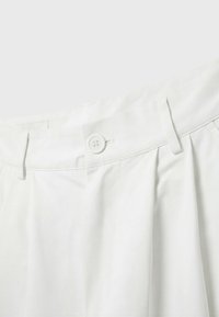 Gros plan sur la taille d’un pantalon blanc avec une fermeture par bouton, des passants de ceinture et des plis sous la taille.