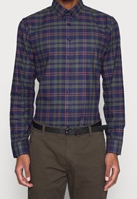 Camicia a scacchi in verde scuro, blu navy e bordeaux; maniche lunghe, colletto button-down, bottoni a contrasto, abbinata a pantaloni scuri e una cintura nera.