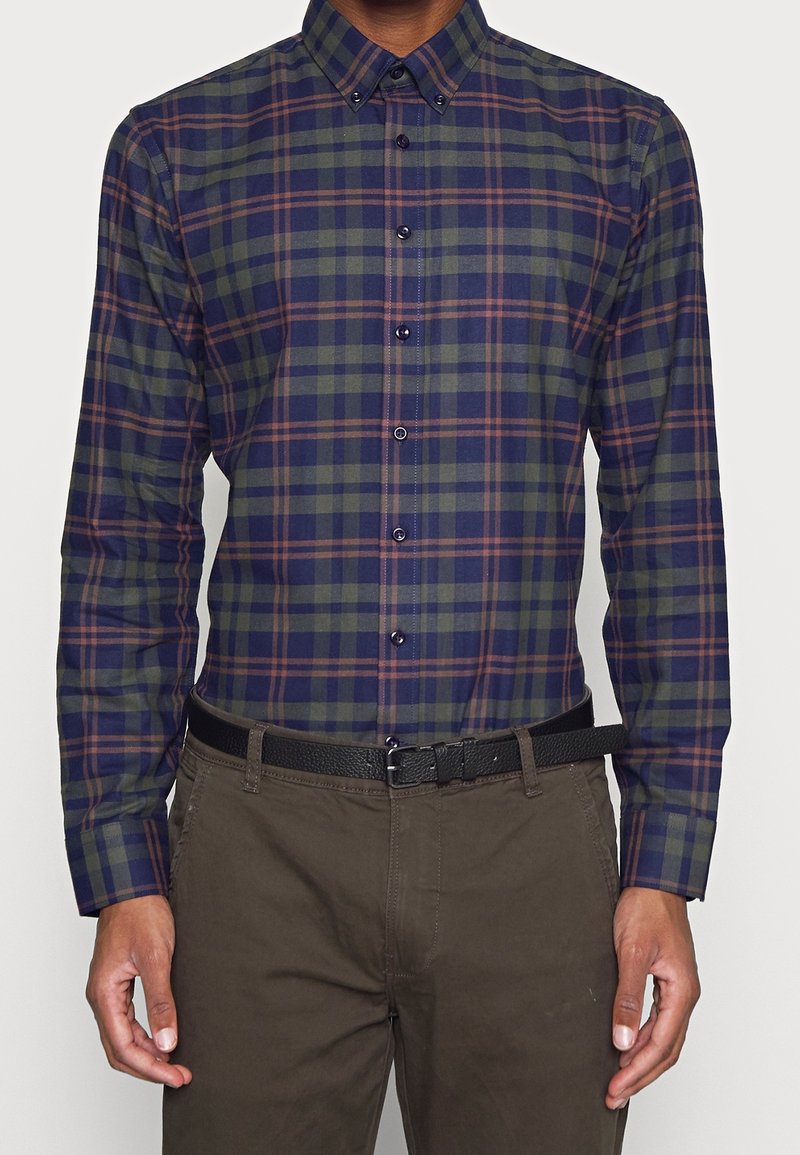 Camicia a scacchi in verde scuro, blu navy e bordeaux; maniche lunghe, colletto button-down, bottoni a contrasto, abbinata a pantaloni scuri e una cintura nera.