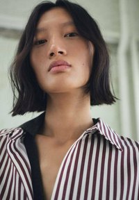 Modelo con cabello oscuro hasta los hombros que lleva una camisa de cuello con finas rayas en color marón y blanco; textura suave y corte relajado.