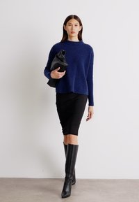 Marineblau, gestrickter Pullover mit lockerer Passform, kombiniert mit einem schwarzen Bleistiftrock und kniehohen schwarzen Lederstiefeln, hält eine schwarze Tasche.
