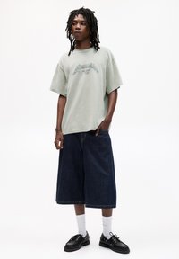 Tricou oversized verde deschis cu un imprimeu grafic, asortat cu culottes din denim albastru închis. Pantofi negri cu șireturi și ciorapi albi completează ținuta.