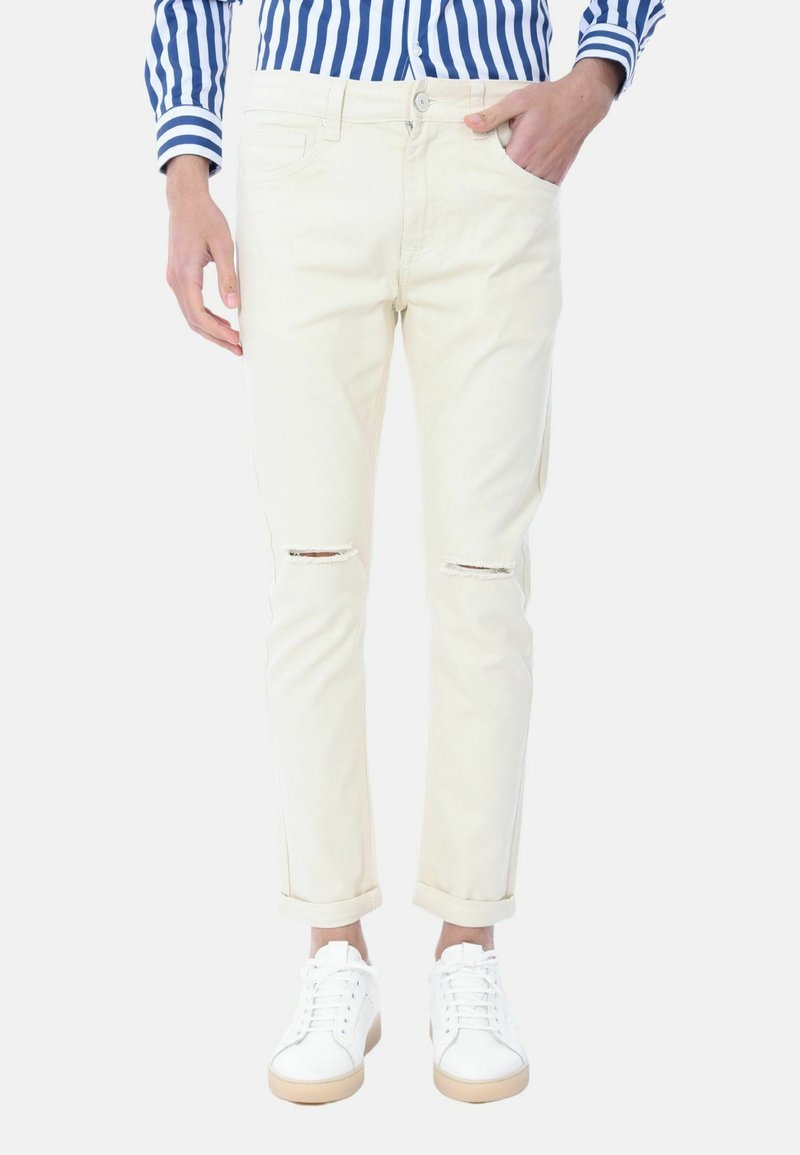 Jeans beige chiaro con effetti vissuti e strappi visibili, orli risvoltati, cinque tasche, abbinati a scarpe da ginnastica bianche. Il modello tiene una mano in tasca.