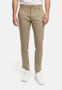 Pantalones beige ajustados hechos de tela suave, con cierre de botón, bolsillos frontales y un diseño de corte slim, combinados con zapatillas blancas.