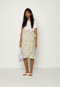 T-shirt branco de mangas curtas combinado com uma saia envolvente floral amarela até aos joelhos, sandálias brancas de plataforma e uma bolsa de ombro lilás clara.