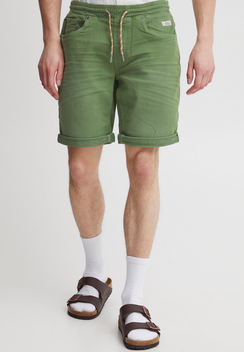 Pantaloncini di denim verdi con risvolti, vita elastica e coulisse. Abbinati a calzini bianchi e sandali marroni.