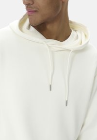 Šviesiai smėlinis hoodie su laisvu kirpimu, turintis dvigubą gobtuvą, raištelius ir sidabro spalvos metalinius antgalius. Lygus paviršius.