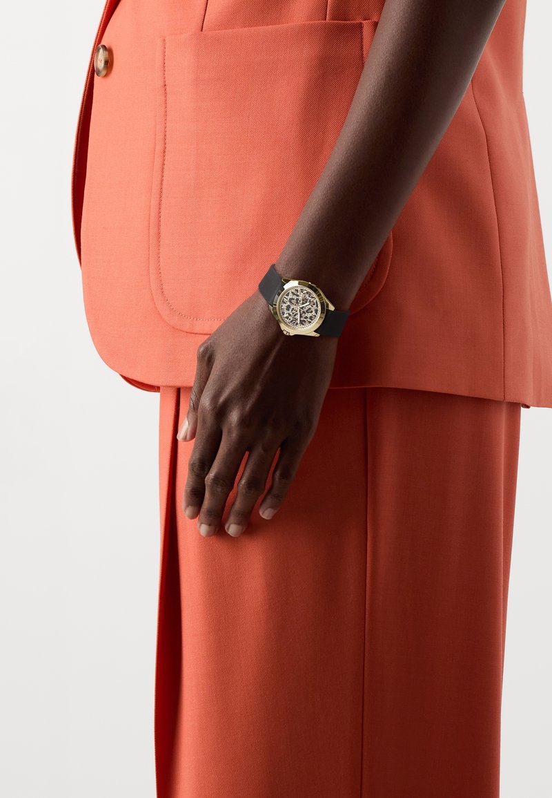 Bras à la peau foncée portant une montre-bracelet à bracelet noir avec un boîtier doré, à côté d’un pantalon orange sur mesure et d’une veste assortie avec un bouton et une poche.